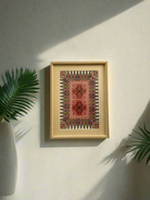 A framed hand-embroidered piece featuring pink and gold colors, displayed on a wall with a shadow cast upon it, alongside a white vase with a green plant.
