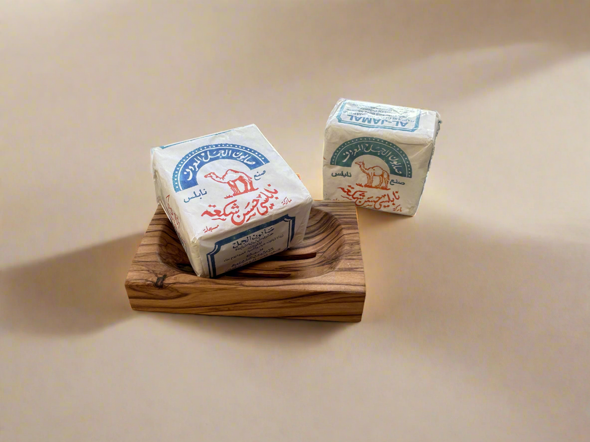 Two bars of olive oil soap on a wooden dish with a shadow falling over the dish, set against a neutral background.