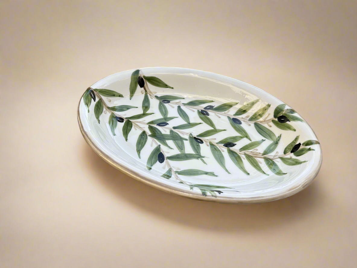 An oval-shaped ceramic serving dish with a handpainted green leaf pattern on a white background, displayed on a neutral backdrop.