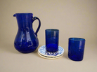 A set of cobalt blue glassware, including a mini pitcher and two tumblers, with a ceramic coaster featuring calligraphy.