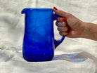 A hand holding a cobalt blue glass pitcher against a white background.
