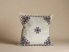 A square-shaped cushion cover with a geometric Canaanite star motif embroidered in purple on a beige background.